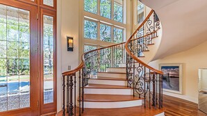Una imponente escalera de caracol con escalones de madera y barandillas de hierro forjado, que conduce a un rellano con un amplio ventanal con vistas a un jardín.