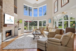 A spacious living room with a fireplace, comfortable seating, large windows, and a view of the outdoors.