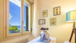 Una habitación con una mesa, una lámpara, obras de arte enmarcadas en la pared y una ventana con vista al mar.