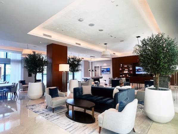 A modern hotel lobby with a seating area, a dining area, and a kitchen in the background.