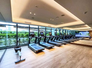 A modern gym with treadmills aligned against a wall of large windows, offering views of greenery and buildings outside. - 2BR Address Dubai Opera | Walk to Mall, Burj Khalifa & Opera (Dubai)