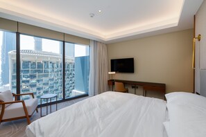 A hotel room with a large bed, a desk, a chair, a TV, and a view of a cityscape through large windows. - 2BR Address Dubai Opera | Walk to Mall, Burj Khalifa & Opera (Dubai)