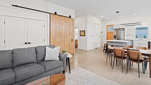 A modern living room with a gray sofa, wooden panel wall, and a dining area with wooden chairs and a table.