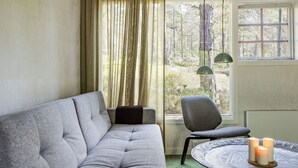 Une pièce chaleureuse avec un canapé gris, une table ronde décorée de bougies et une vue sur une forêt, visible à travers une fenêtre ornée de rideaux.