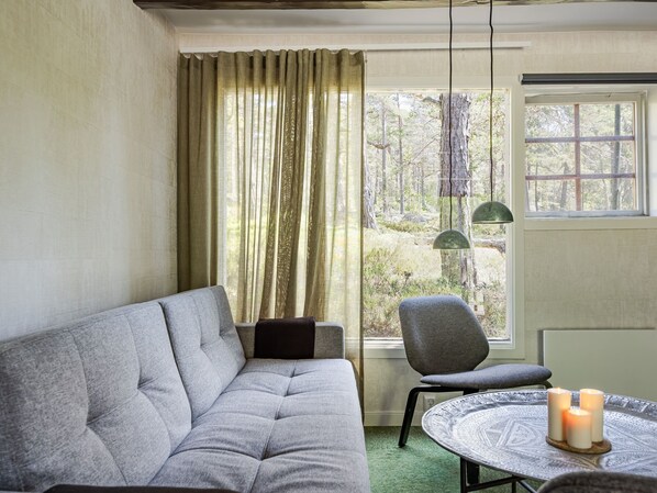 A cozy room with a grey sofa, a round table with candles, and a view of a forest through a window with curtains.