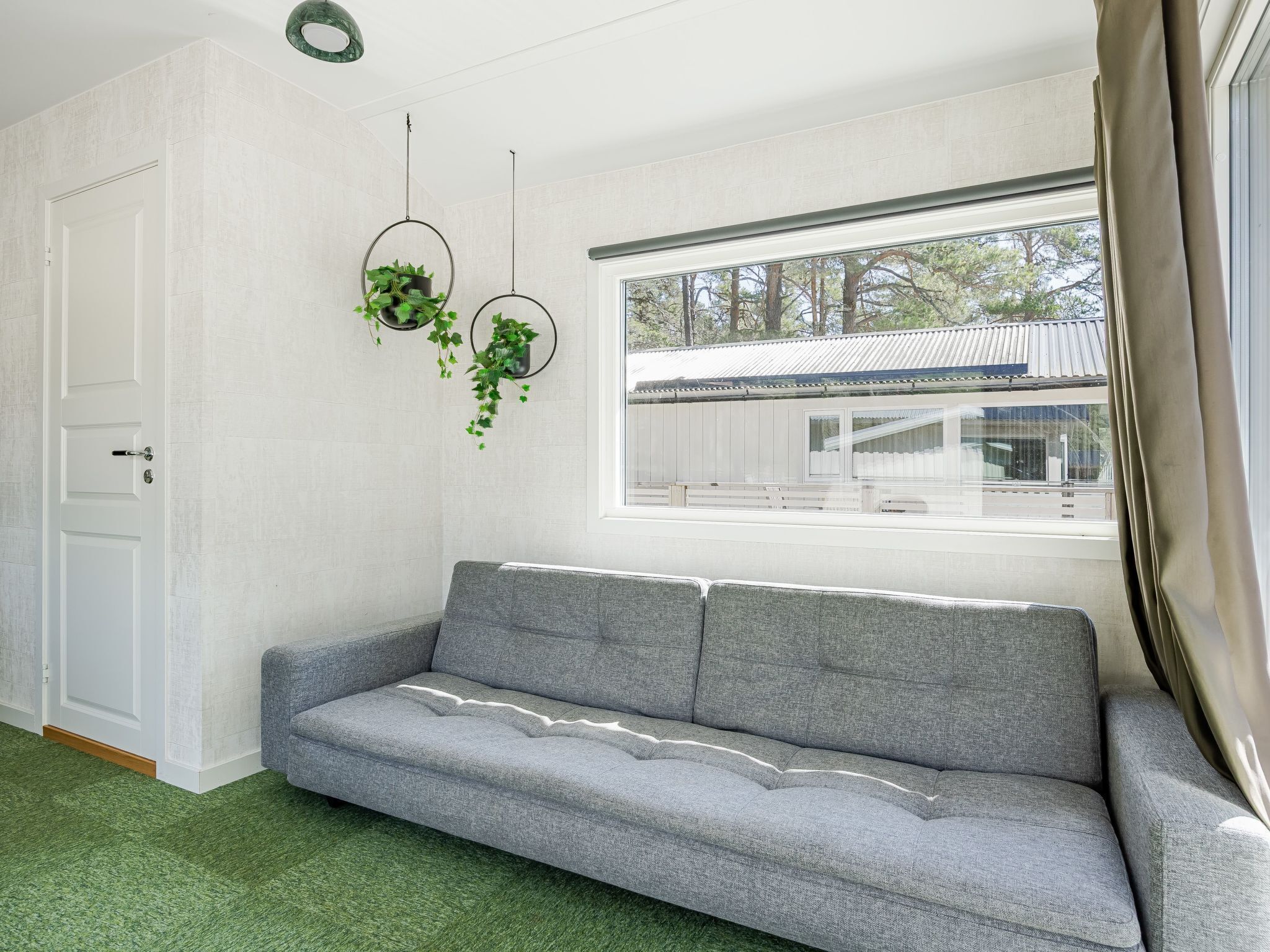 A room with a grey sofa, a white door, and a window with a view of trees and a building.