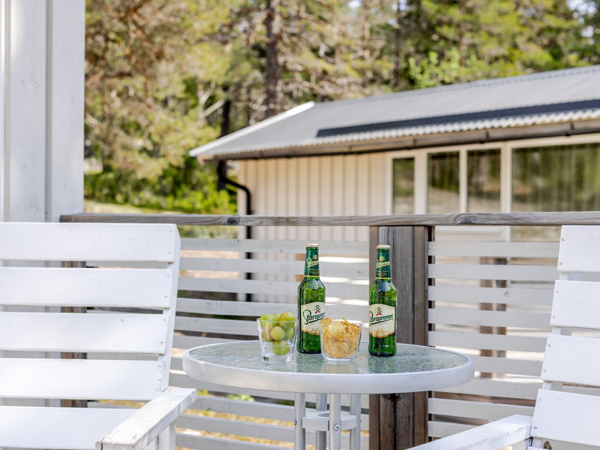 Een ronde tafel met twee Carlsberg biertjes, een glas groene druiven en een schaal chips op een balkon met witte houten stoelen.