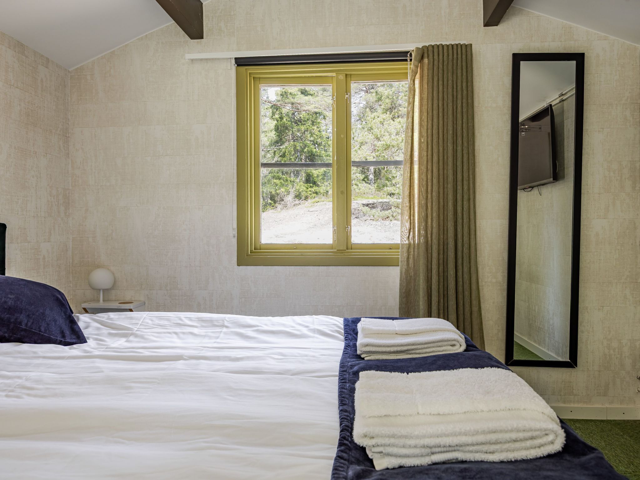 A bedroom with a bed, a window with curtains, a bedside table with a lamp, and a mirror on the wall.
