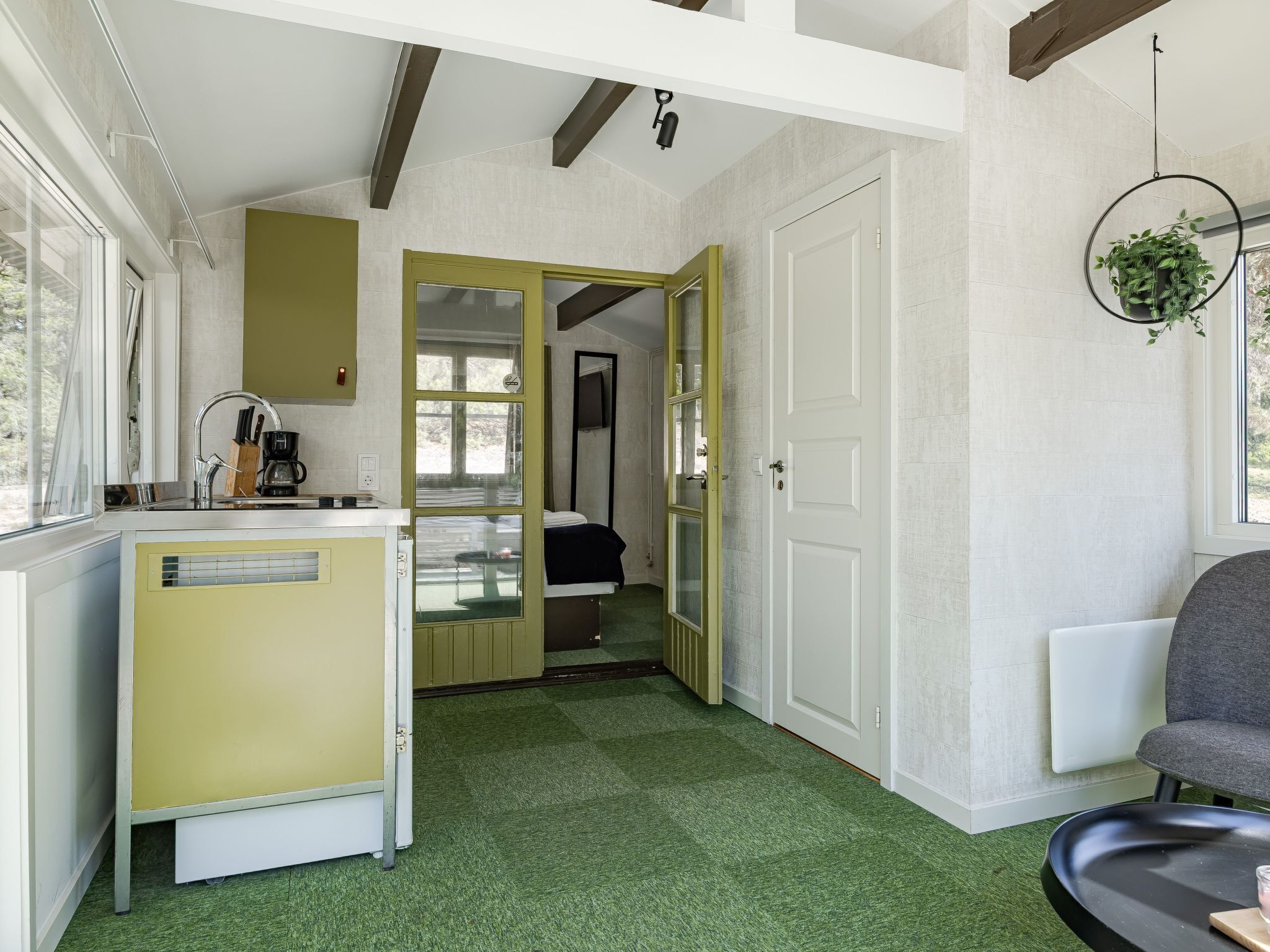 A compact kitchen with a sink, a coffee maker, and a window. There is a bed with a green headboard and a door leading to another room. A chair is visible in the foreground.