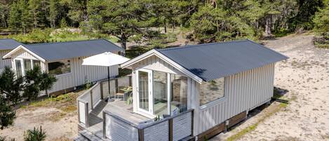 Une petite cabane à un seul étage, dotée d’un balcon et entourée d’arbres et de sable.