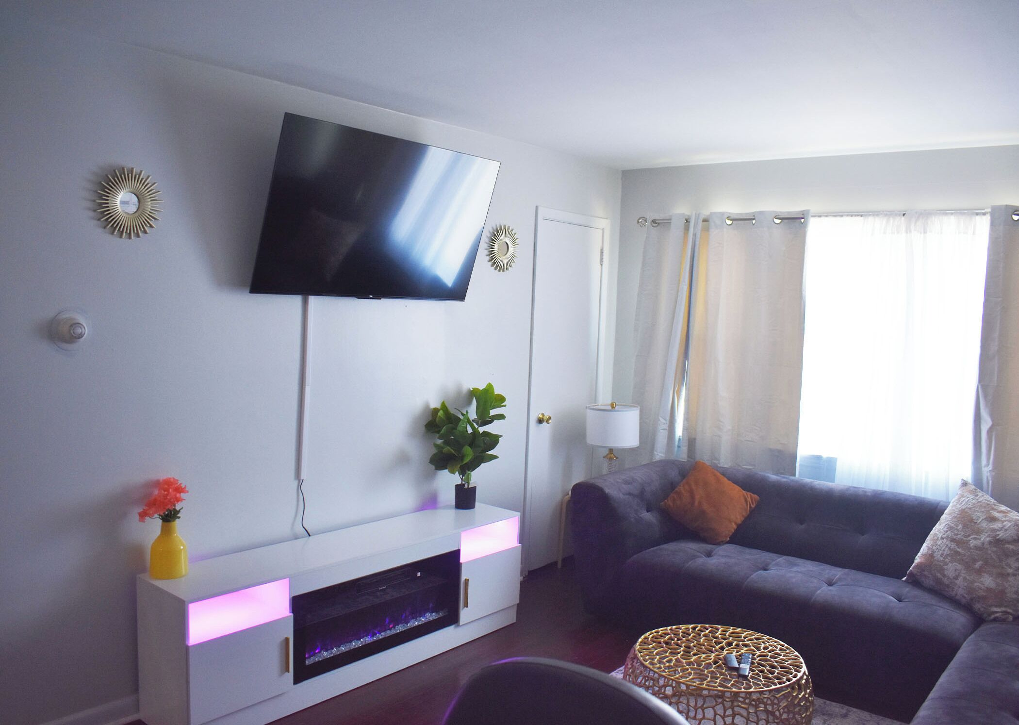 En stue med et fladskærms-tv på væggen, et hvidt tv-møbel med en plante og en vase, en mørk sofa med puder, et lille rundt bord og et vindue med gardiner.