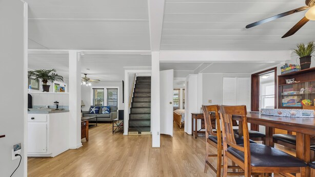 A spacious living area with a wooden floor, a dining table with chairs, a staircase, and a kitchen area with white cabinets and a countertop.