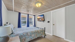 A bedroom with a bed, a nightstand, a lamp, a TV, and a view of the outside.