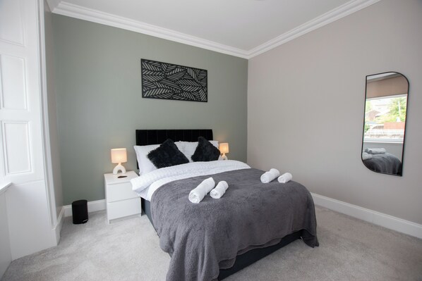 A neatly made bed with a grey blanket and pillows, a bedside table with a lamp, and a wall-mounted mirror.