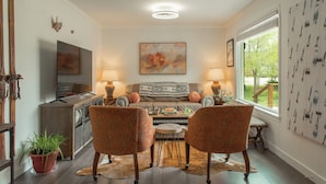 A cozy living room with a sofa, two patterned chairs, a coffee table, and a TV. There is a window with a view of greenery, a fireplace, and a potted plant. - Heber base camp, sleeps 7, fire pit, pets ok, AC (Heber City)