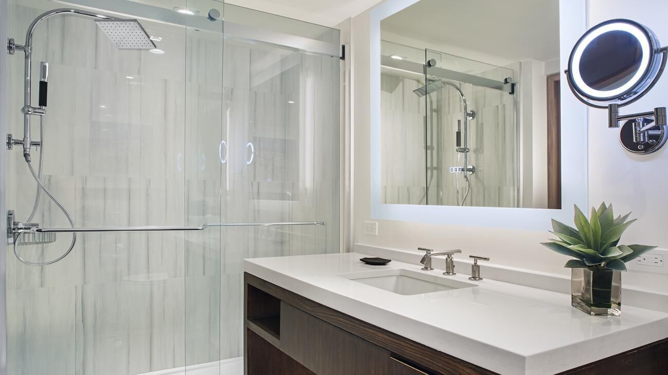 Une salle de bain moderne avec une douche à l’italienne en verre, un grand miroir avec un luminaire rond et une plante en pot posée sur le meuble-vasque.