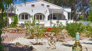 Una villa blanca con ventanas arqueadas y un balcón, rodeada por un jardín con una estatua y una botella de vino sobre una mesa.