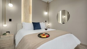 A modern hotel room with a neatly made bed, a bedside table with a plant, and a tray of fruit and cheese.