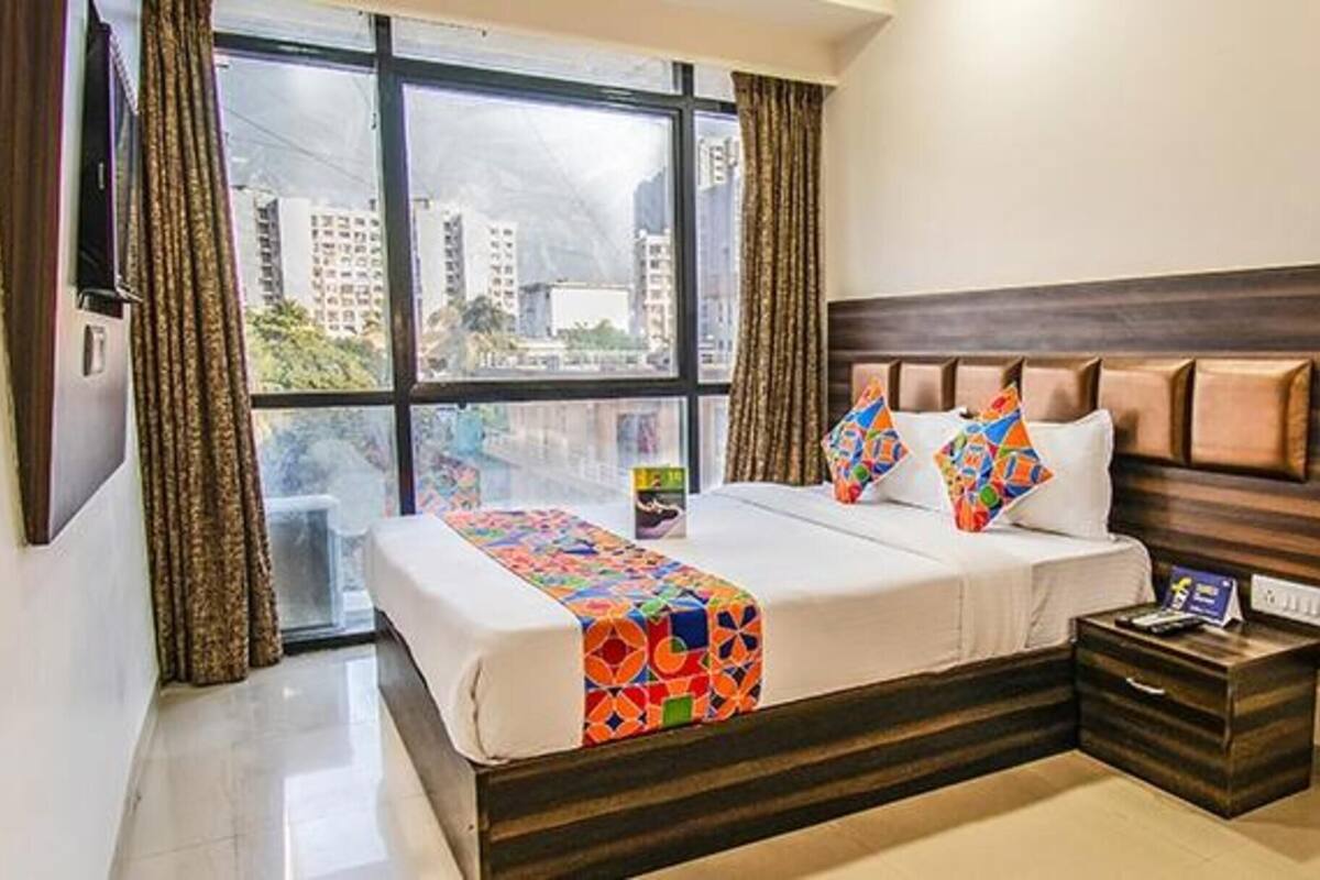 A hotel room with a large window, a bed with colorful bedding, a wooden headboard, and a nightstand with a remote control.