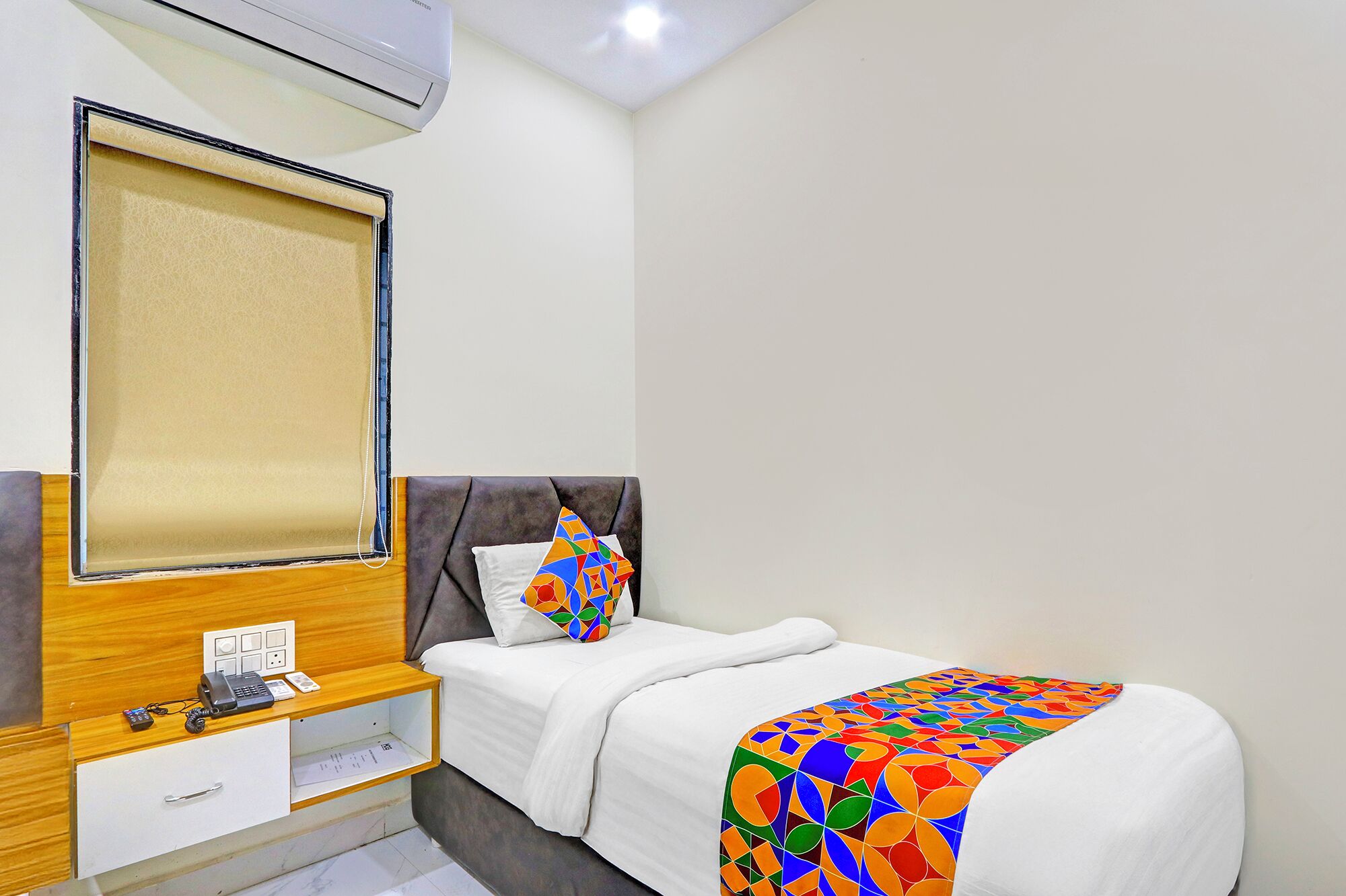 Une chambre d’hôtel avec un lit, une table de chevet avec un téléphone et une lampe, un miroir et une fenêtre avec des stores.