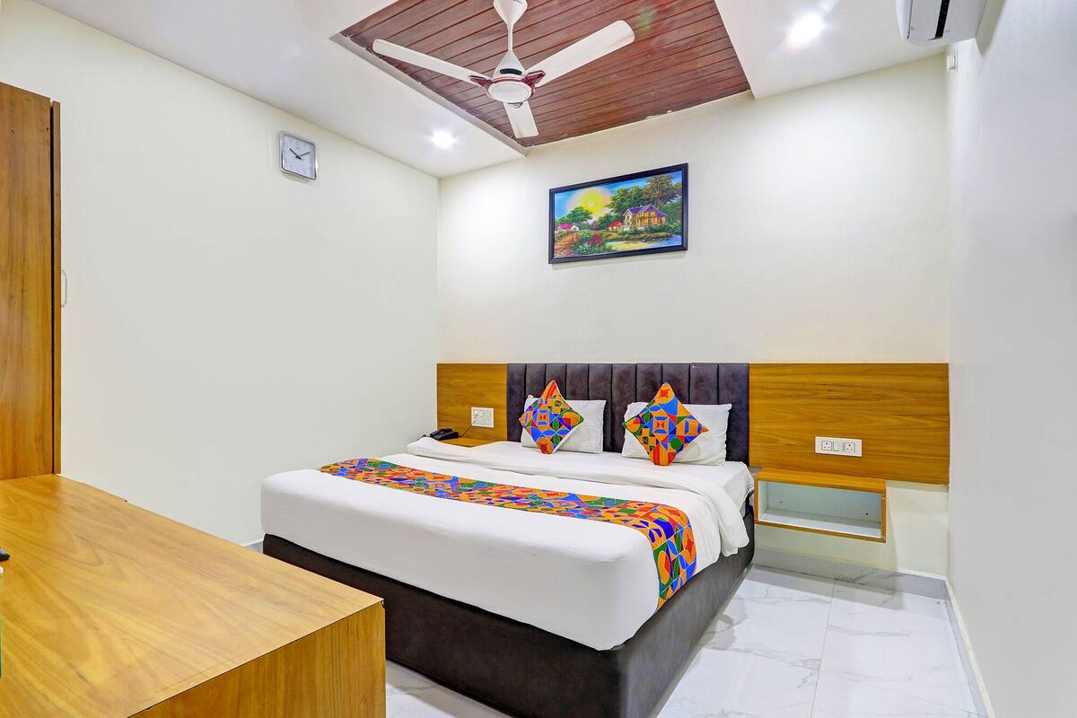 A hotel room with a bed, a wooden headboard, a ceiling fan, a painting on the wall, and a small table.