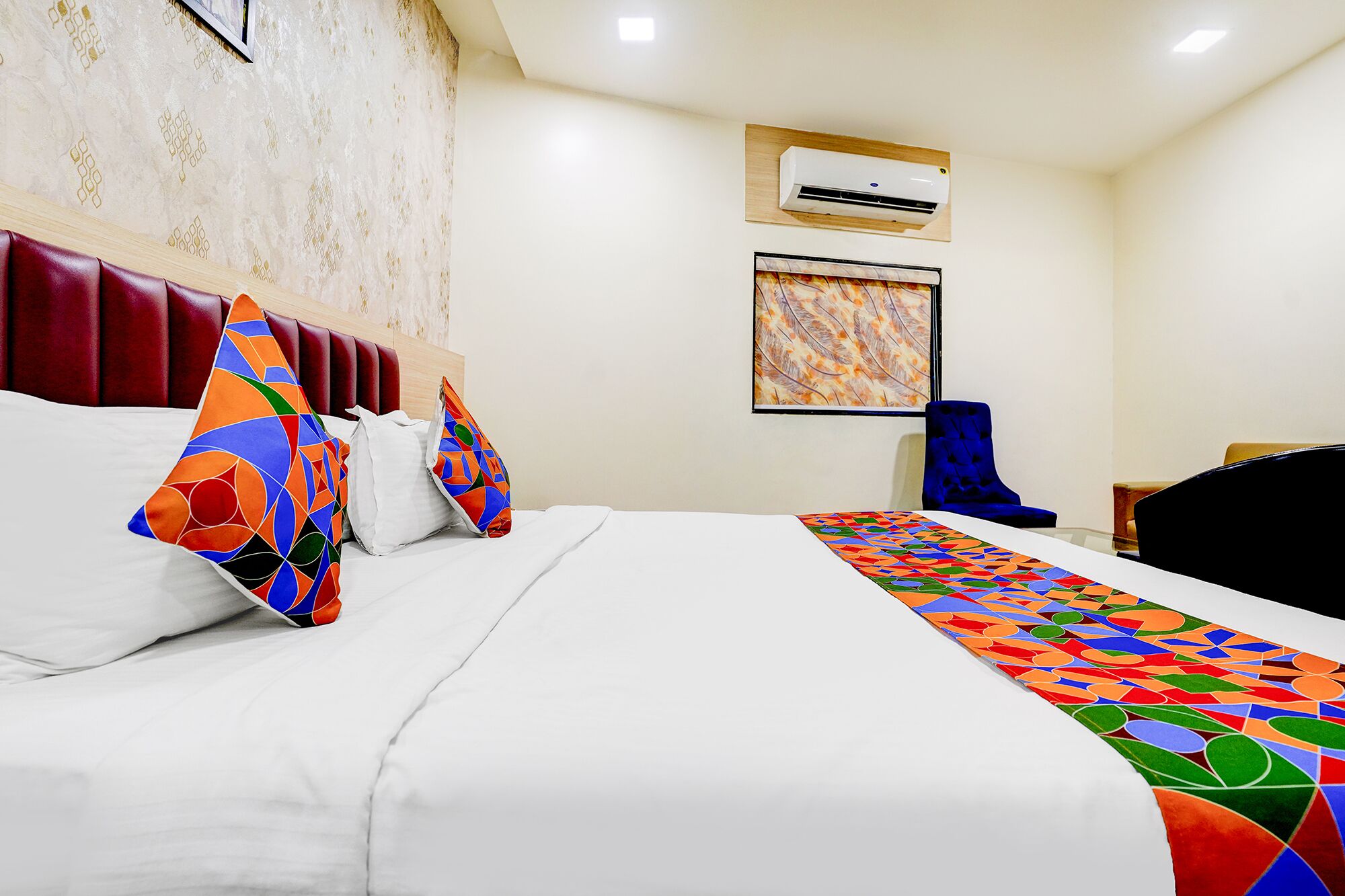 A hotel room with a bed, a headboard, an air conditioning unit, a painting, a blue chair, and a desk.