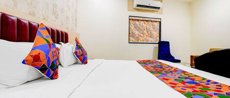 A hotel room with a bed, a headboard, an air conditioning unit, a painting, a blue chair, and a desk.