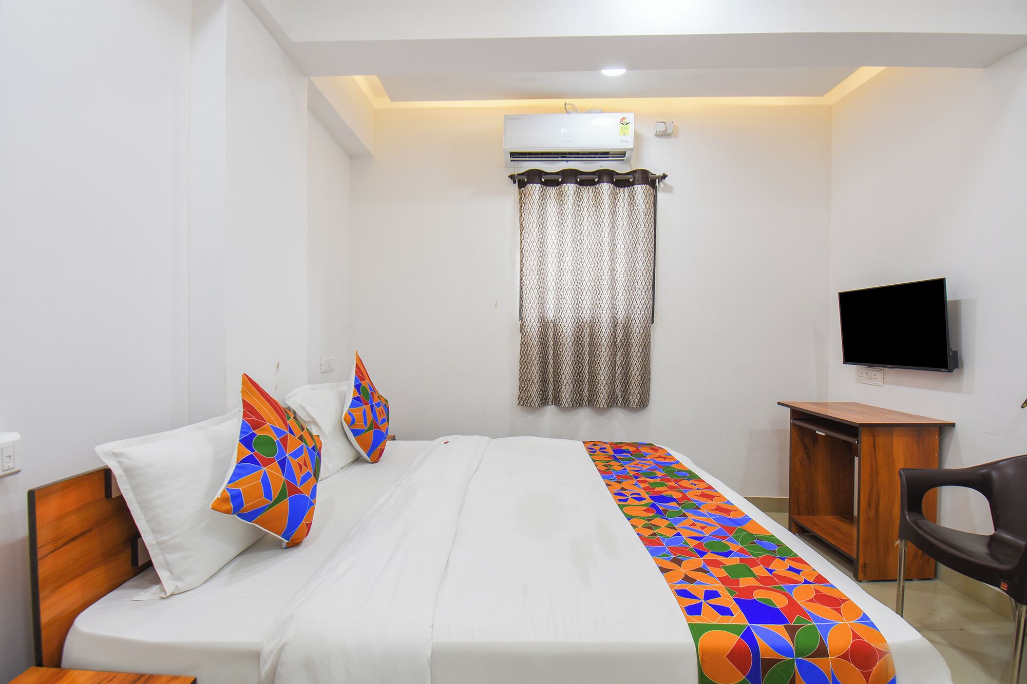 A hotel room with a bed, a desk, a chair, an air conditioner, a TV, and a window with curtains.