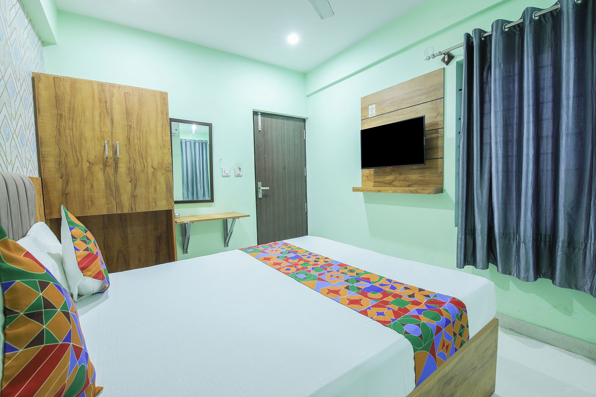 A bedroom with a bed, a wooden wardrobe, a small desk, a mirror, a TV mounted on the wall, and a window with curtains.
