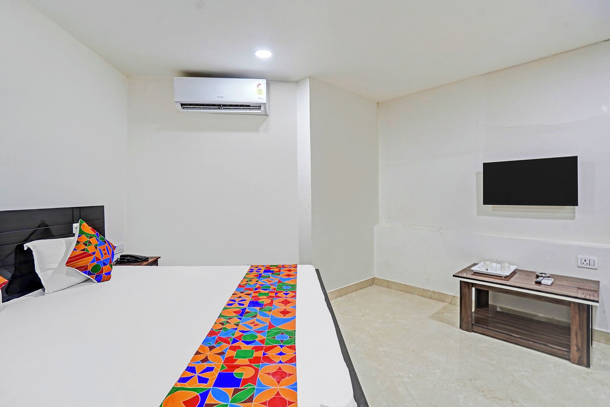 A hotel room with a bed, a bedside table with a lamp, a TV mounted on the wall, and an air conditioning unit.