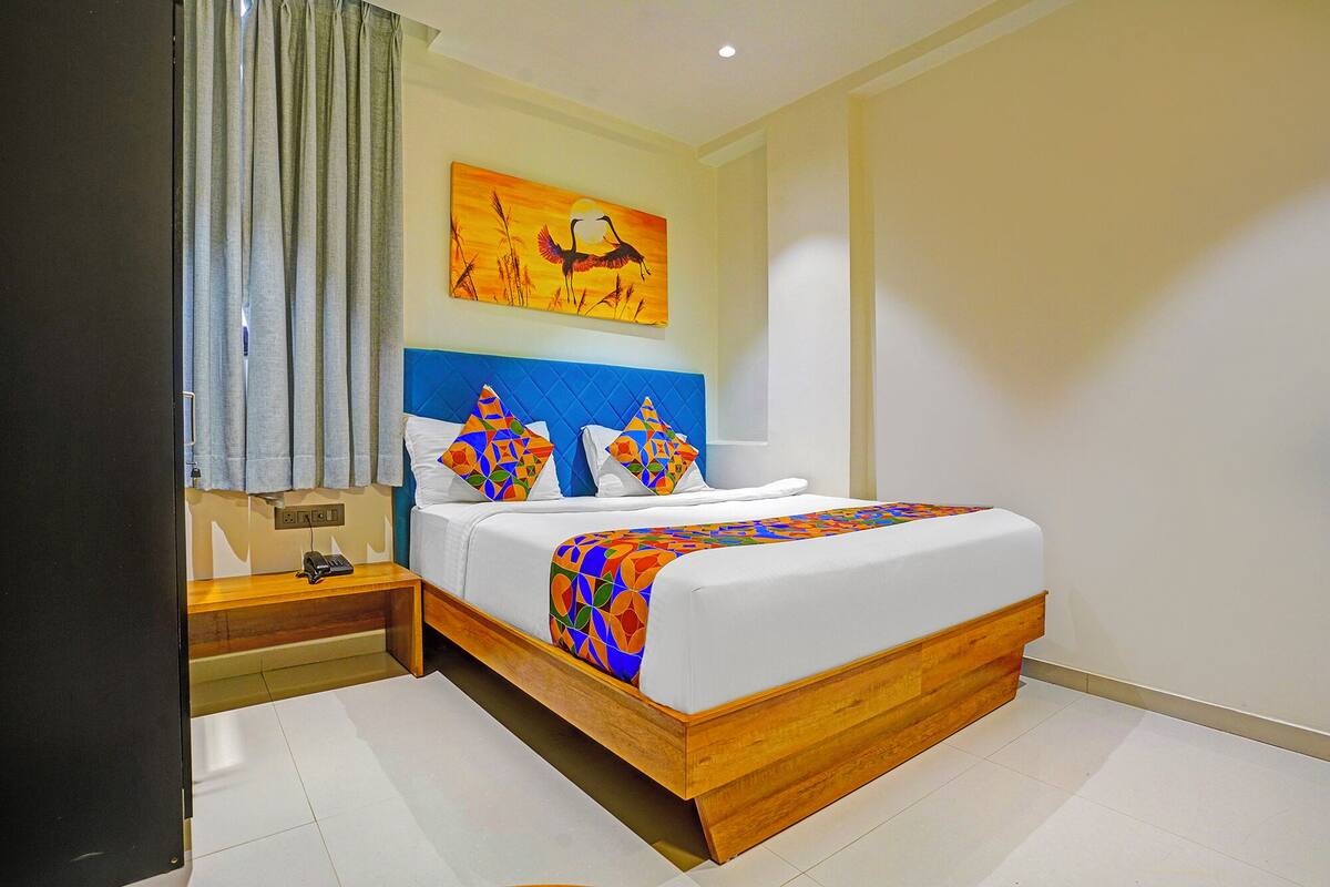 Quarto de hotel com cama, mesa de cabeceira, quadro e janela com cortinas.