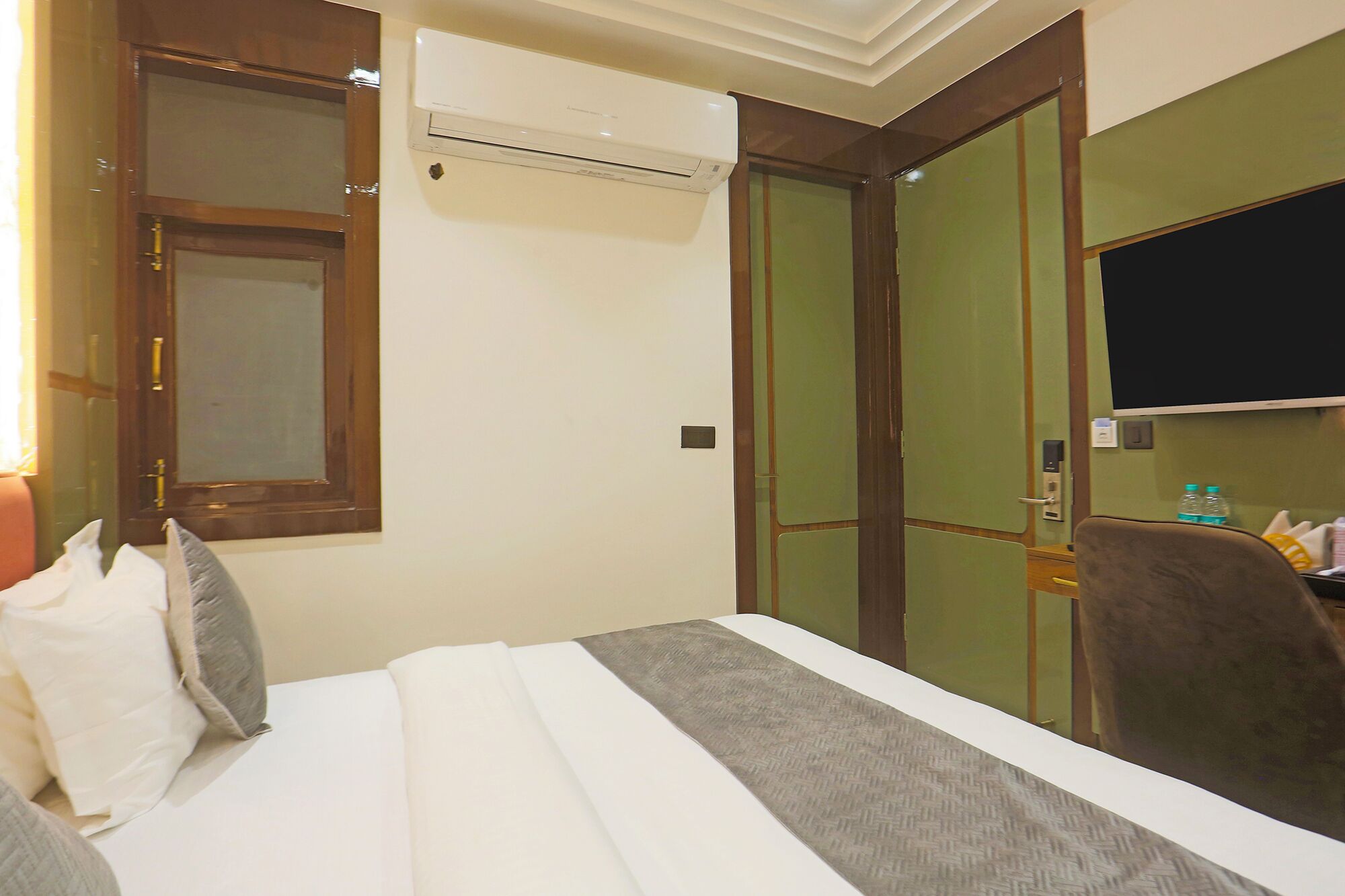 A hotel room with a bed, a window, an air conditioning unit, a television, and a door with a green panel.