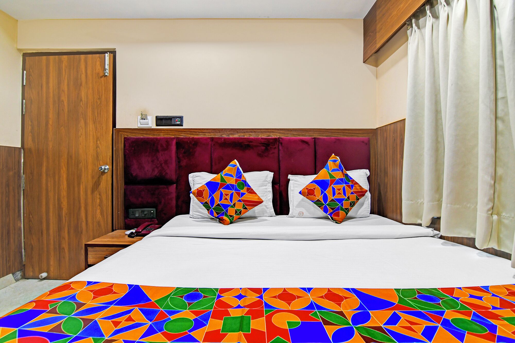 A hotel room with a wooden door, a red upholstered headboard, a bed with white bedding, two colorful geometric pillows, and a patterned bedspread.