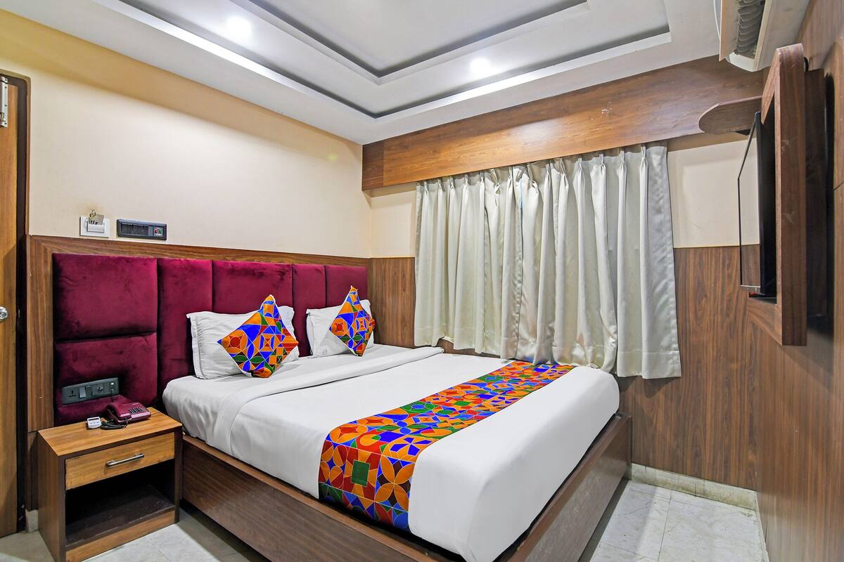 A hotel room with a wooden headboard, a bed with white linens, a colorful geometric throw blanket, a bedside table with a telephone, and a window with white curtains.