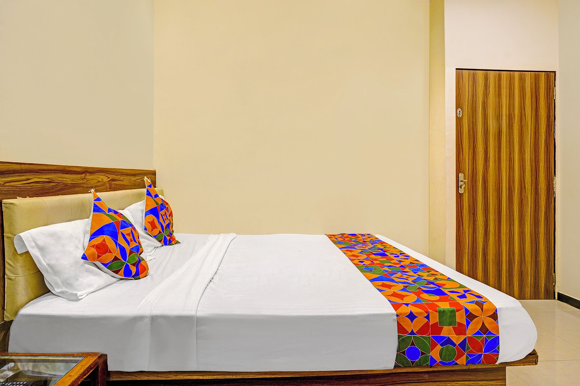 A neatly made bed with a white bedspread and colorful geometric pillows, a wooden headboard, and a wooden door in the background.