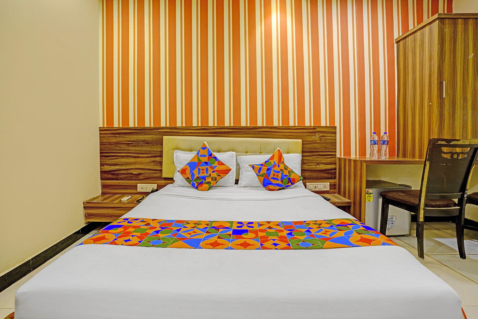 A hotel room with a bed, two pillows, a nightstand, a chair, a wooden headboard, and a striped wall.