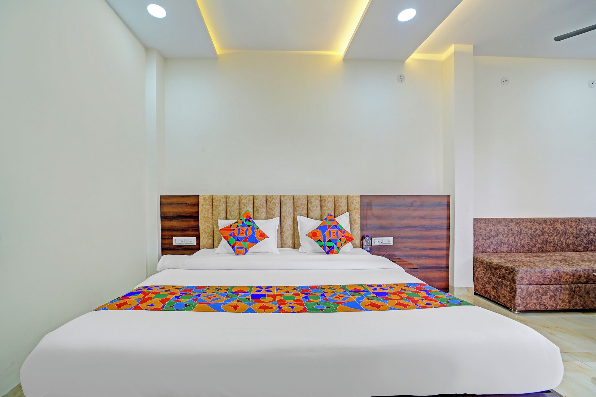 A hotel room with a large bed, a headboard with a geometric pattern, a colorful bedspread, and a brown upholstered chair.