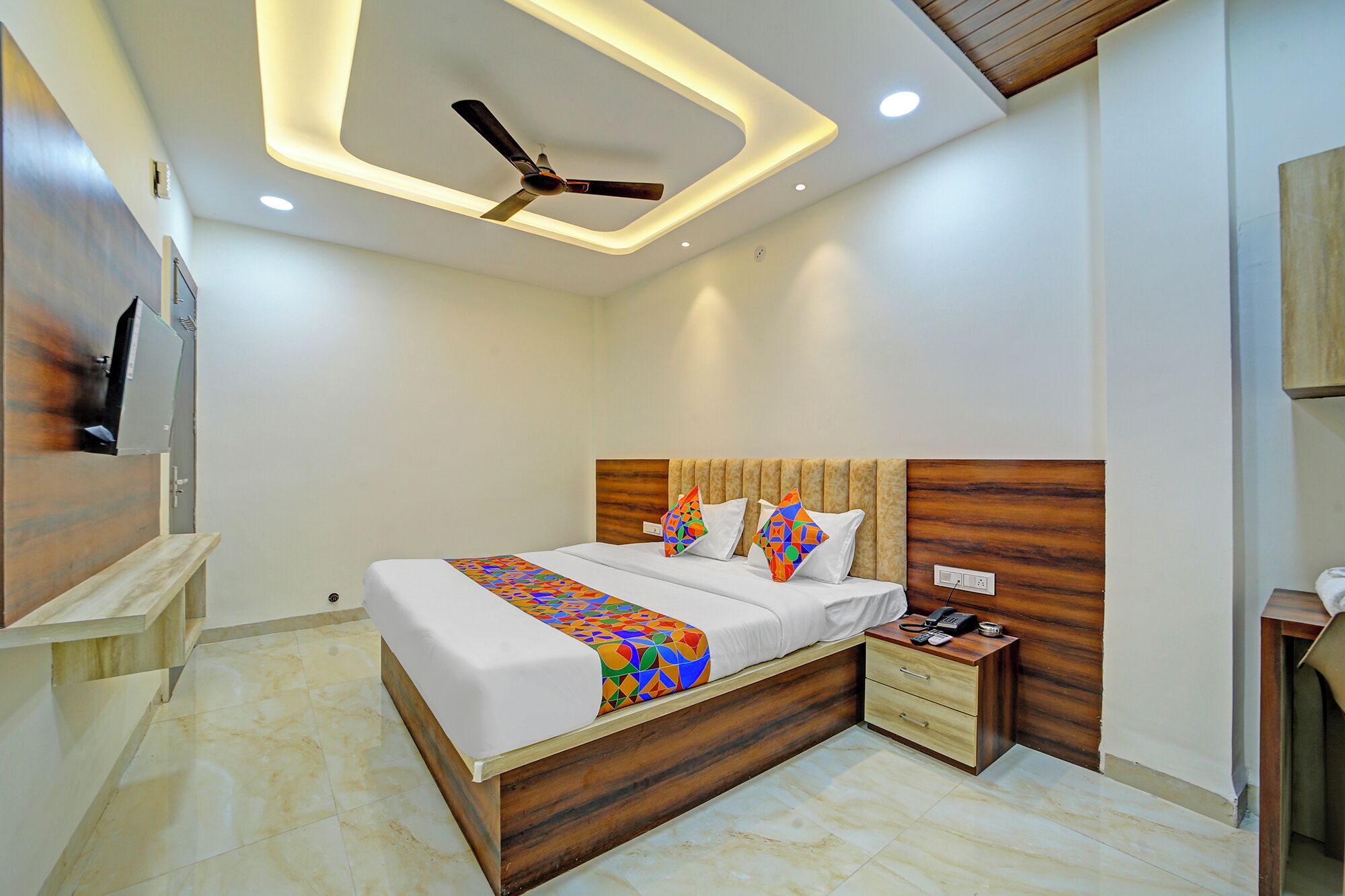 Une chambre moderne avec une tête de lit en bois, un lit agrémenté d’une literie aux motifs géométriques colorés, un ventilateur de plafond et une table de chevet sur laquelle se trouvent un téléphone et un chargeur.