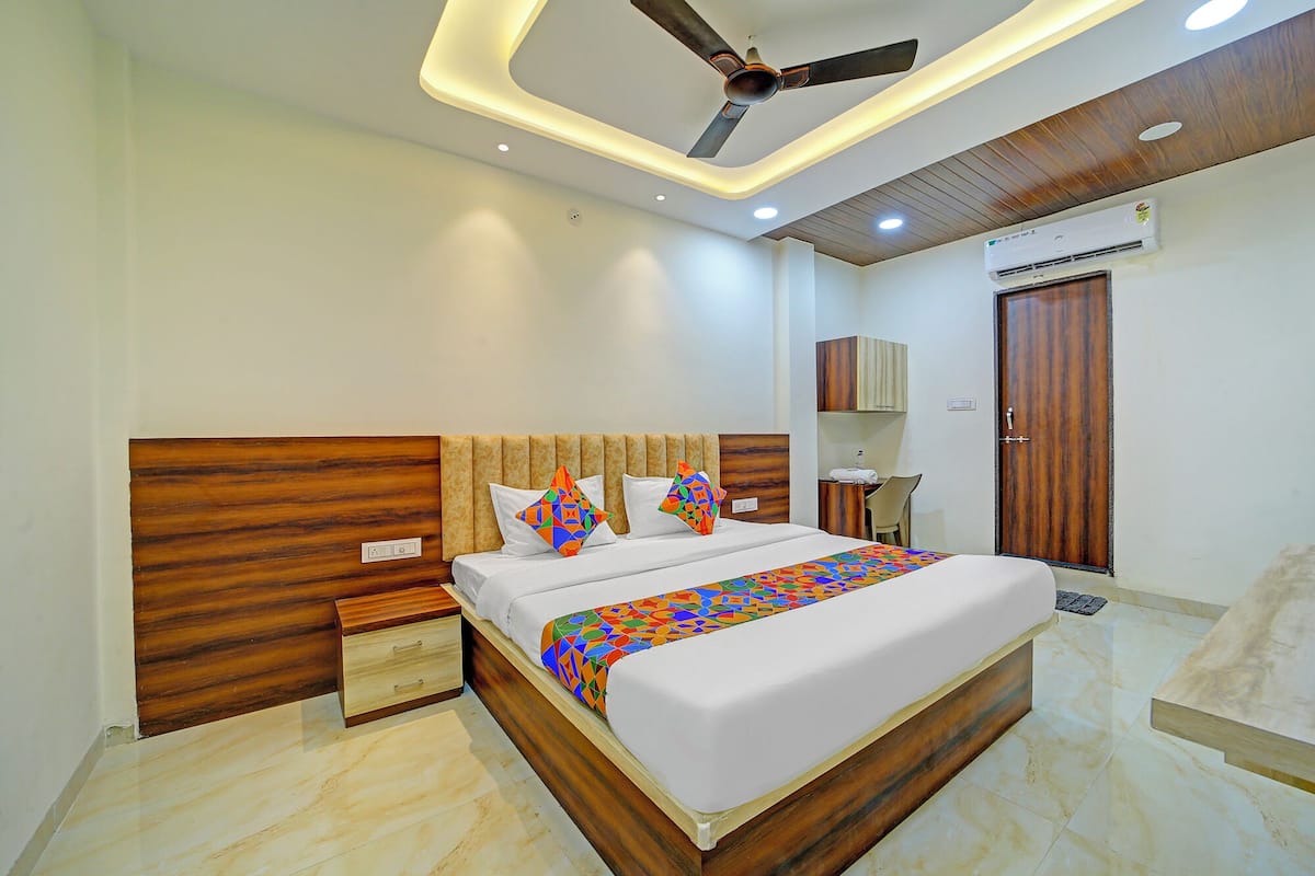 A modern hotel room with a wooden headboard, a ceiling fan, a bed with colorful bedding, a bedside table, a wardrobe, and an air conditioning unit.