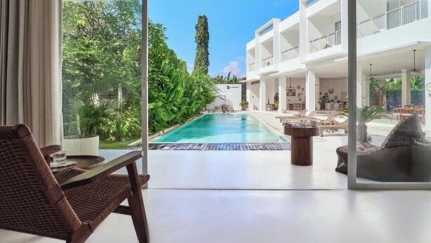 A modern hotel room with a large glass door leading to a pool area, surrounded by lush greenery and a multi-level building. - Serendipity Legian (Legian)