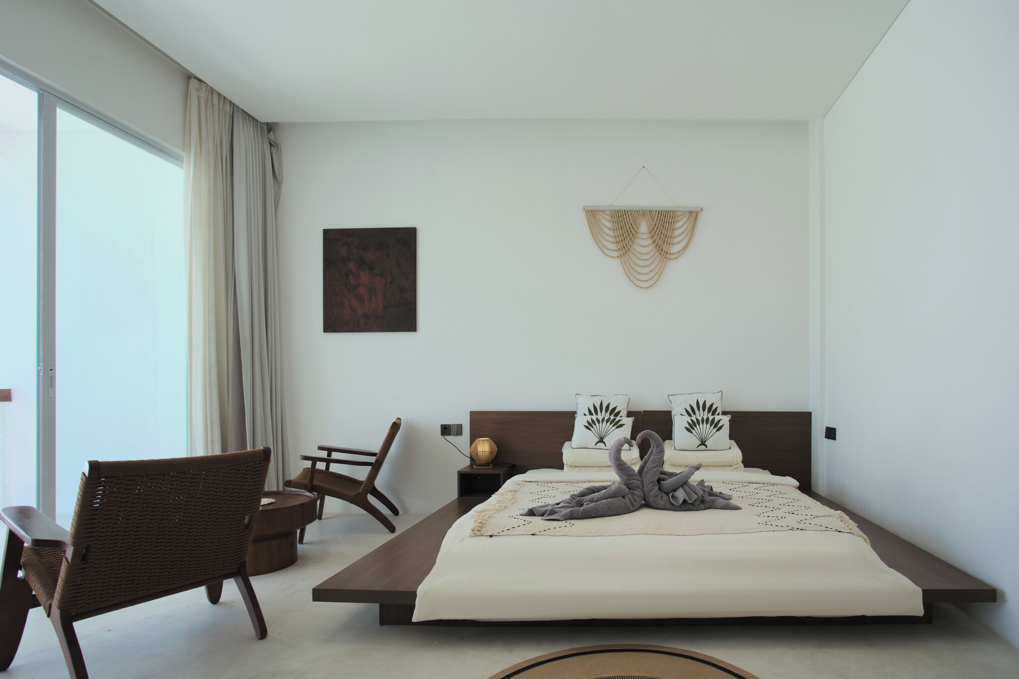 Un dormitorio moderno con una cama grande, dos mesitas de noche, una silla de mimbre y vistas al exterior a través de una puerta corredera.