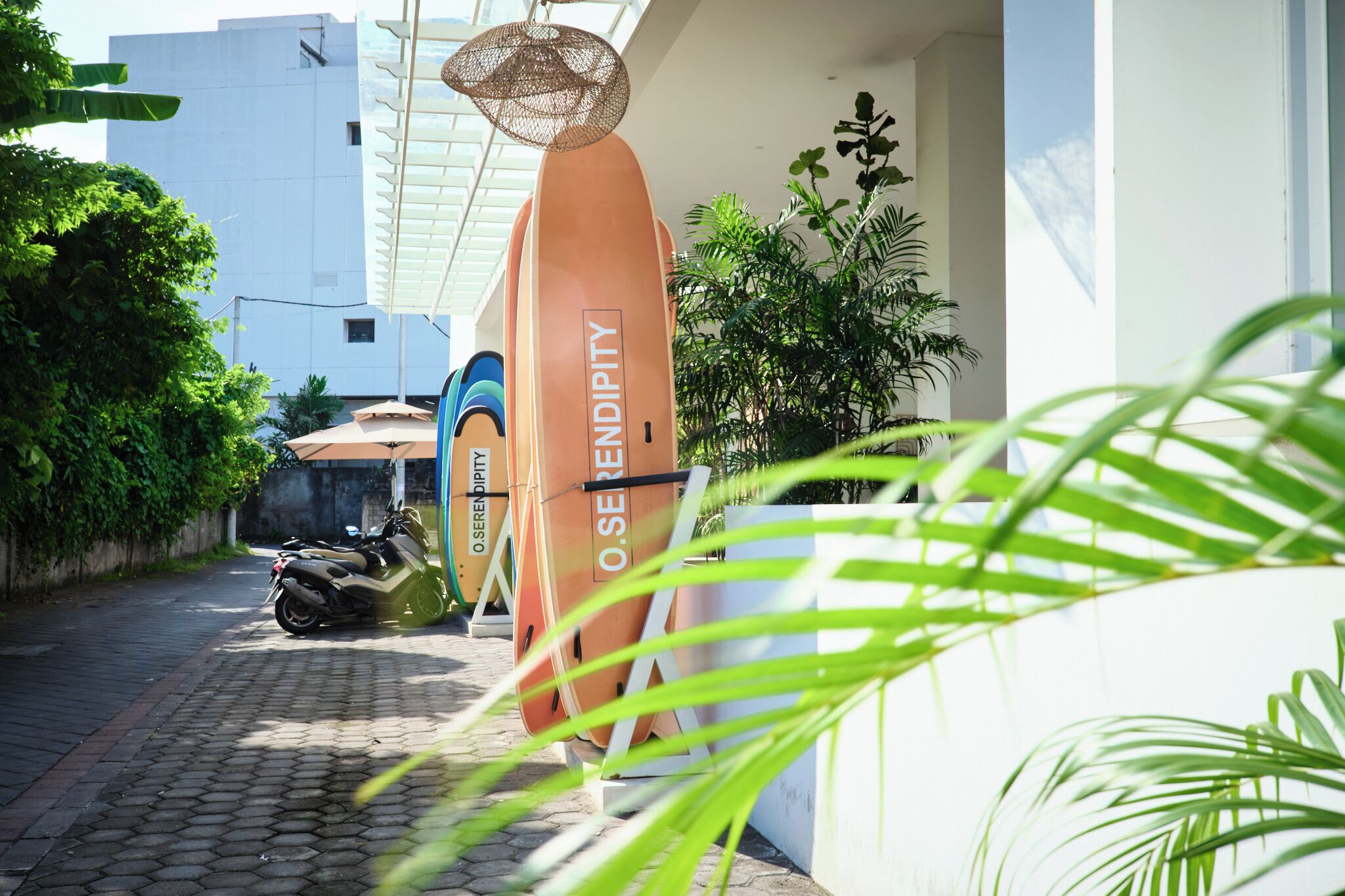 A narrow alley with a parked motorcycle, a large orange sculpture labeled "OSEREN DIPITY," and a building with a white facade and green plants.