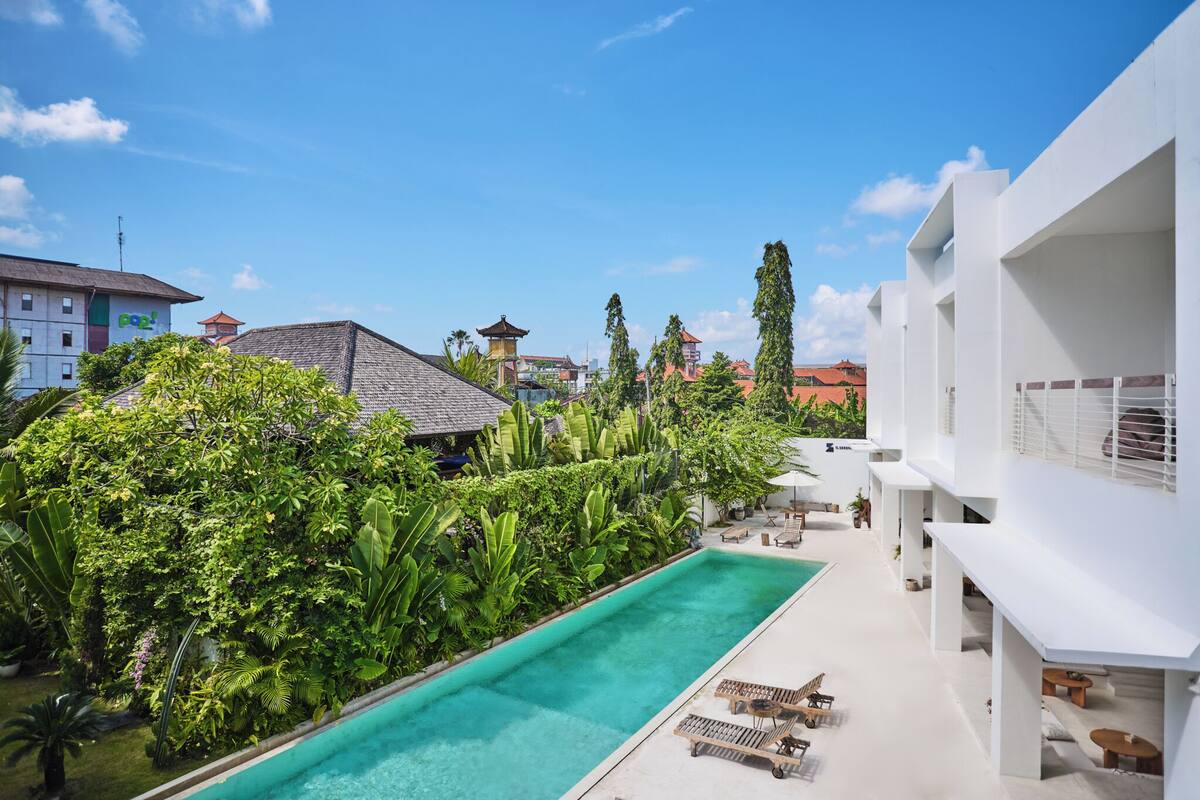 A swimming pool surrounded by lush greenery, a white building with balconies, and traditional architecture in the background.