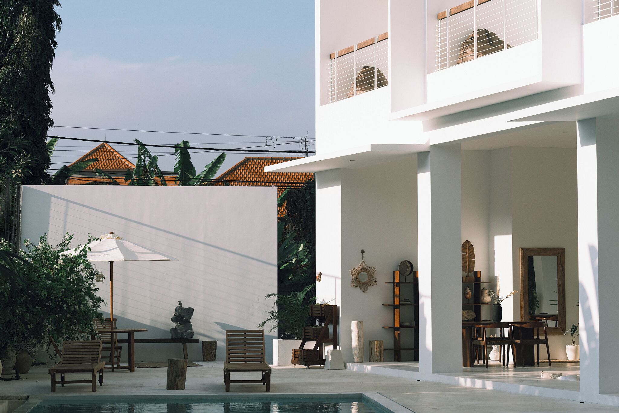 A modern outdoor area with a pool, wooden furniture, and a white building with a tiled roof in the background.