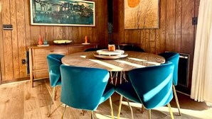 Un coin repas avec une table ronde, des chaises bleues, des murs en bois et deux tableaux.