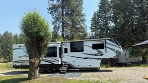 Un grand camping-car doté d’un espace de vie couvert, d’une échelle et d’une table de pique-nique, situé dans une zone herbeuse bordée d’arbres.