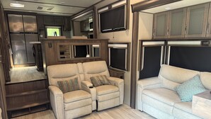 Un intérieur de camping-car spacieux comprenant un coin salon, une cuisine équipée d’un réfrigérateur et d’un four à micro-ondes, et un téléviseur fixé au-dessus du coin salon.