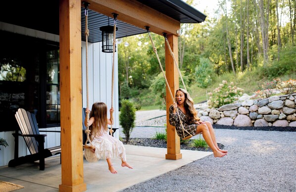 A wooden swing set in a covered outdoor area with a stone wall and greenery in the background. - Private Barndo- Sleeps 18, Hot Tub, Sports Barn, 30 Min to Notre Dame (Cassopolis)