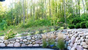Un jardín con un muro de piedra, diversas plantas y un bosque al fondo.