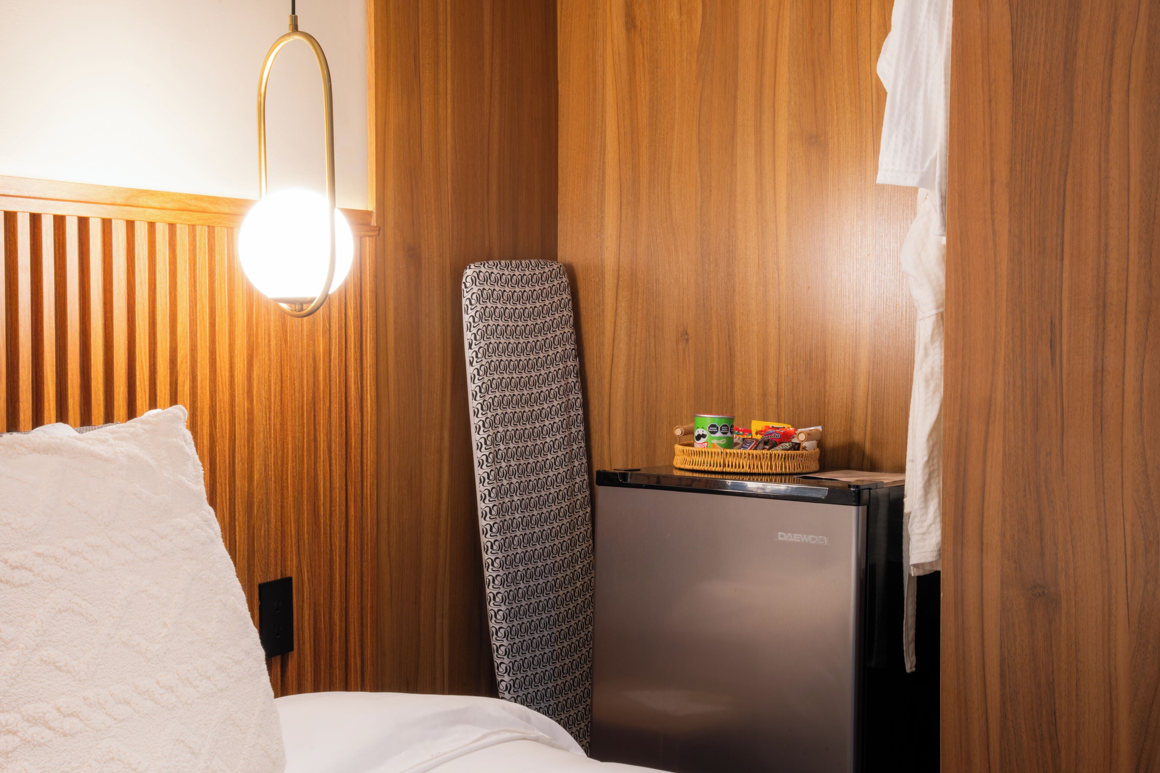 A compact room with a bed, a small refrigerator, a wall-mounted ironing board, and a hanging light fixture.